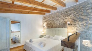 A bedroom with a stone wall, wooden beams, a large bed, bedside tables with lamps, and a wardrobe. - Agritourism SA Franquesa Vella (Vilafranca de Bonany)
