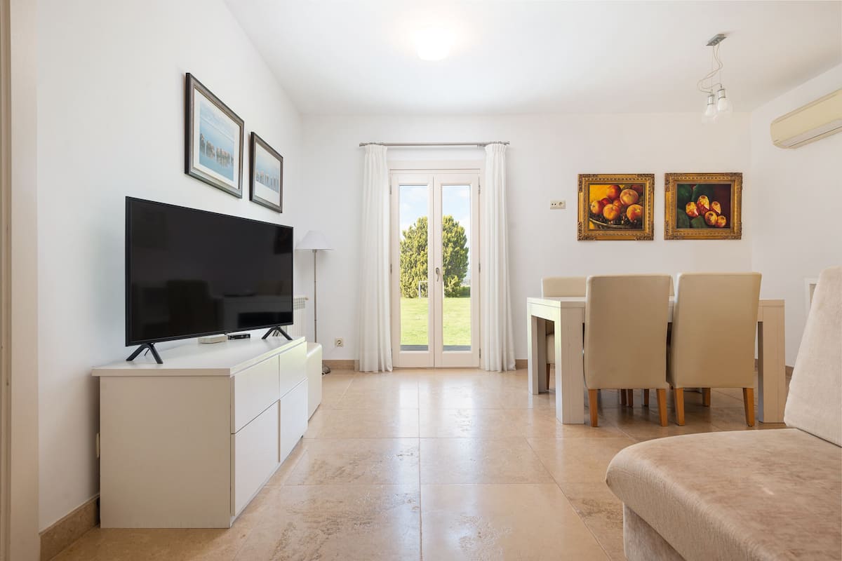 En moderne stue med et fladskærms-tv, en sofa, et spiseområde med stole og en skydedør af glas, der fører ud til en have.