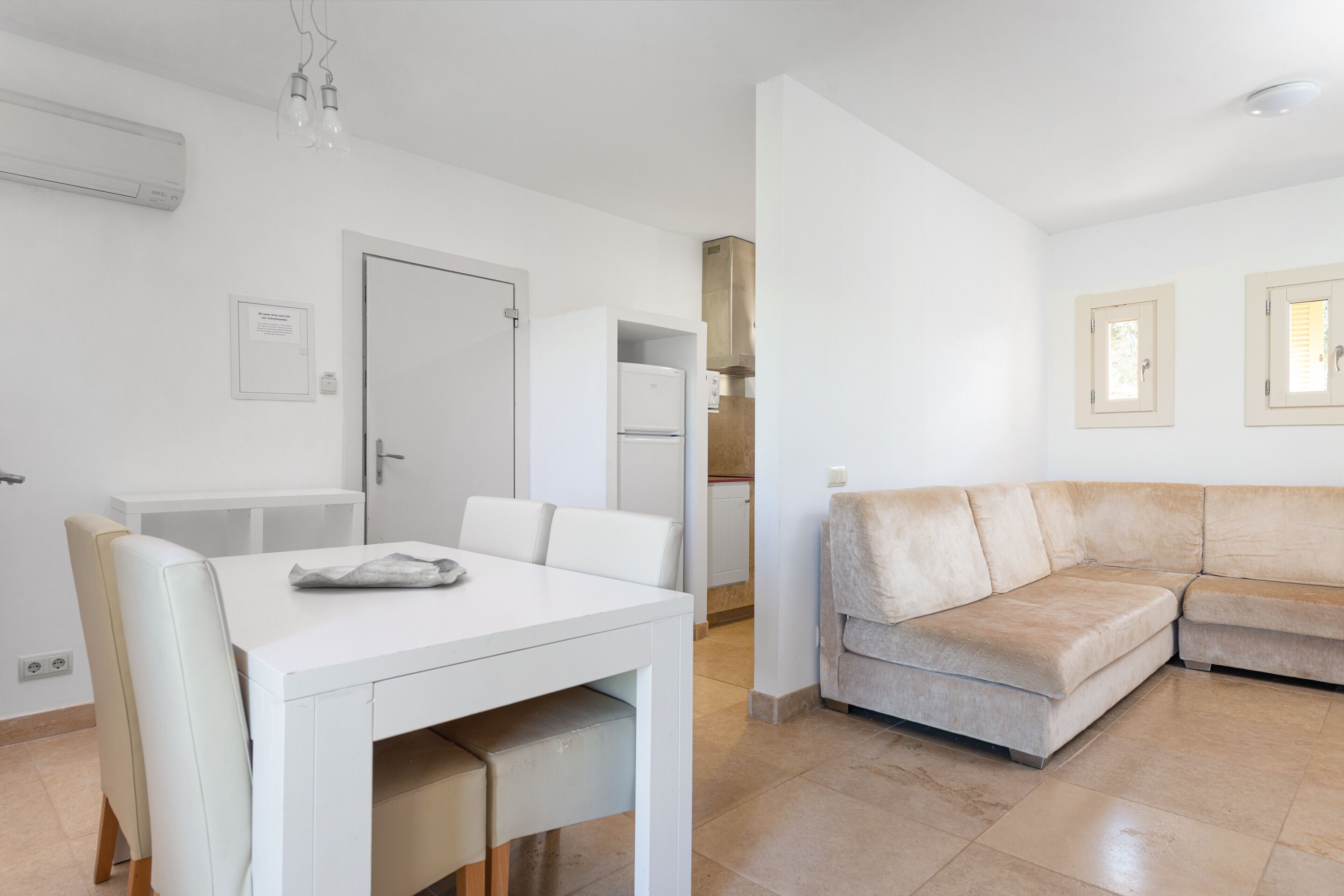 Una sala de estar moderna con un sofá seccional color beige, una mesa de comedor blanca con sillas, y un área de cocina con refrigerador y armarios.