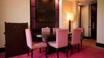 A hotel room with a round table, six patterned chairs, a flat-screen TV, a standing lamp, and a minibar.