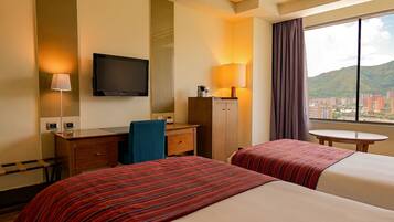 A hotel room with two beds, a desk, a television, and a window with a view of buildings and mountains.