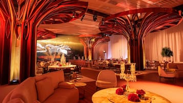 A spacious hotel lobby with a modern design, featuring a central seating area with sofas and a round table, surrounded by multiple dining tables set for guests.