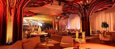 Un amplio hall de hotel con diseño moderno, que presenta una zona de estar central con sofás y una mesa redonda, rodeada de varias mesas de comedor preparadas para los huéspedes.