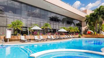 A swimming pool with lounge chairs and umbrellas, surrounded by a building with large windows and tropical plants.