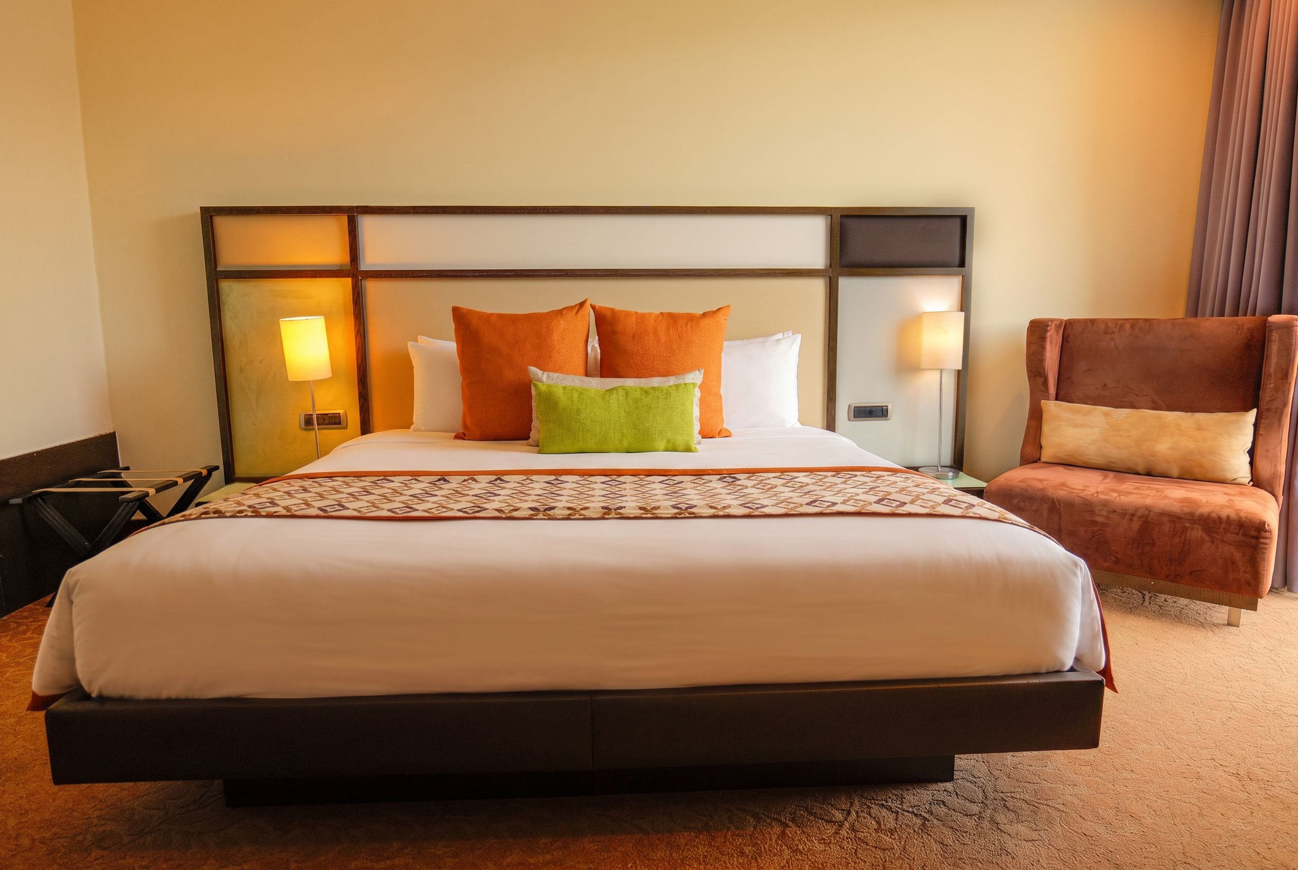Ein Hotelzimmer mit einem großen Bett, zwei Nachttischlampen, einem braunen Sessel und einem gemusterten Teppich.
