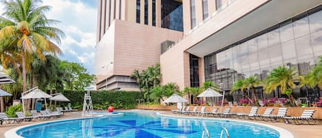 Una piscina con sillas de descanso y sombrillas, rodeada de palmeras y un edificio moderno.