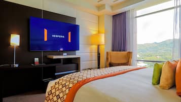 Une chambre d’hôtel dotée d’un grand téléviseur à écran plat fixé au mur, d’un lit avec une literie à motifs, d’un bureau avec une lampe et offrant une vue sur un paysage montagneux par la fenêtre.