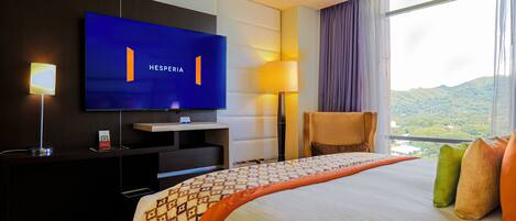Habitación de hotel con un televisor de pantalla plana grande montado en la pared, una cama con ropa de cama estampada, un escritorio con lámpara y vistas a un paisaje montañoso a través de la ventana.
