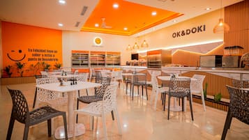 L’intérieur d’un restaurant, avec des tables et des chaises, un comptoir où sont exposés des plats, et un mur orné d’un visage souriant et d’un texte.