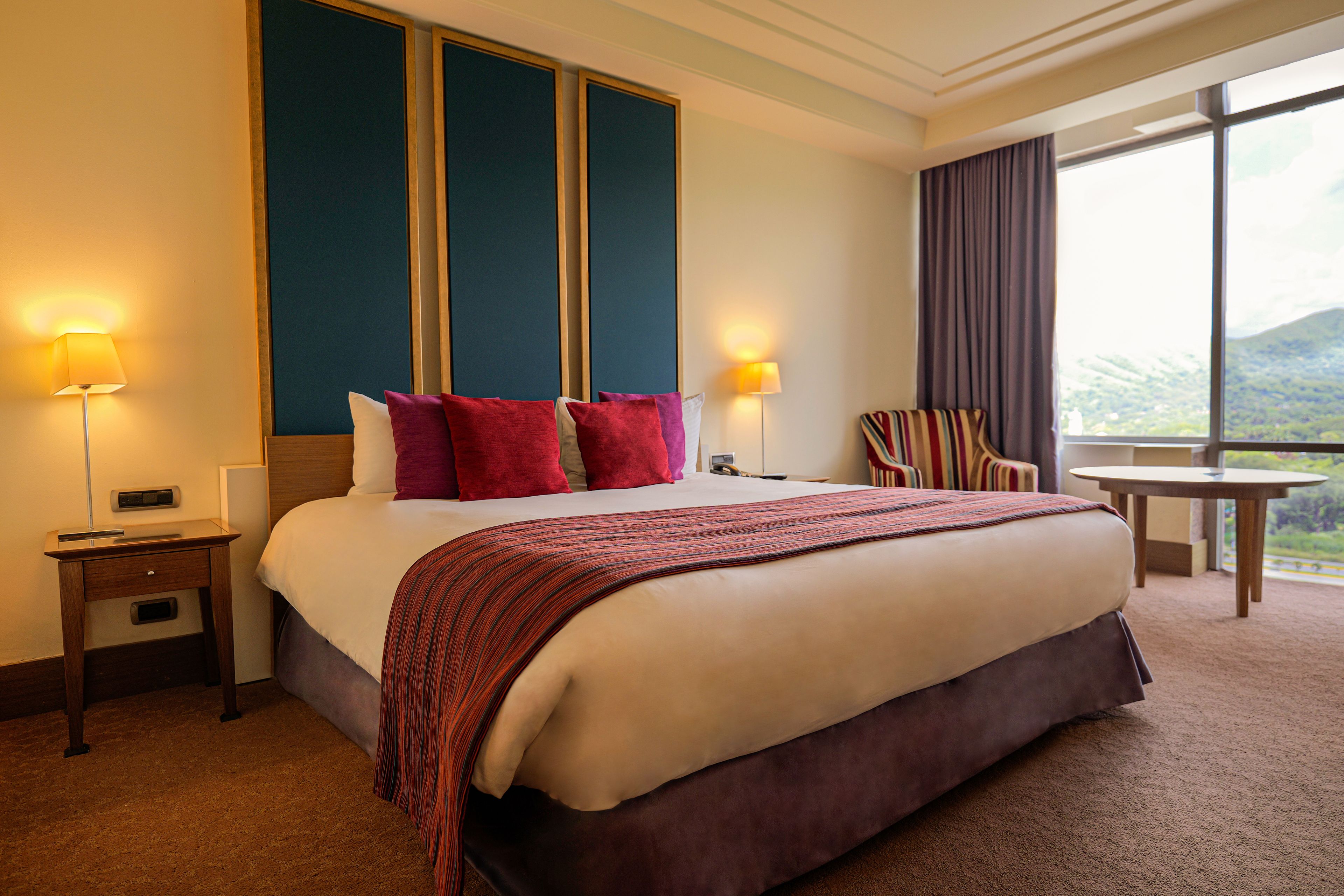 A hotel room with a large bed, two bedside lamps, a nightstand, a chair, a small table, and a window with a view.