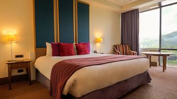 Ein Hotelzimmer mit einem großen Bett, zwei Nachttischlampen, einem Nachttisch, einem Sessel, einem kleinen Tisch und einem Fenster mit Blick.