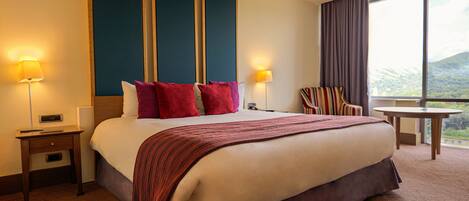 A hotel room with a large bed, two bedside lamps, a nightstand, a chair, a small table, and a window with a view.