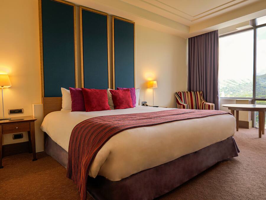 Ein Hotelzimmer mit einem großen Bett, zwei Nachttischlampen, einem Nachttisch, einem Sessel, einem kleinen Tisch und einem Fenster mit Blick.