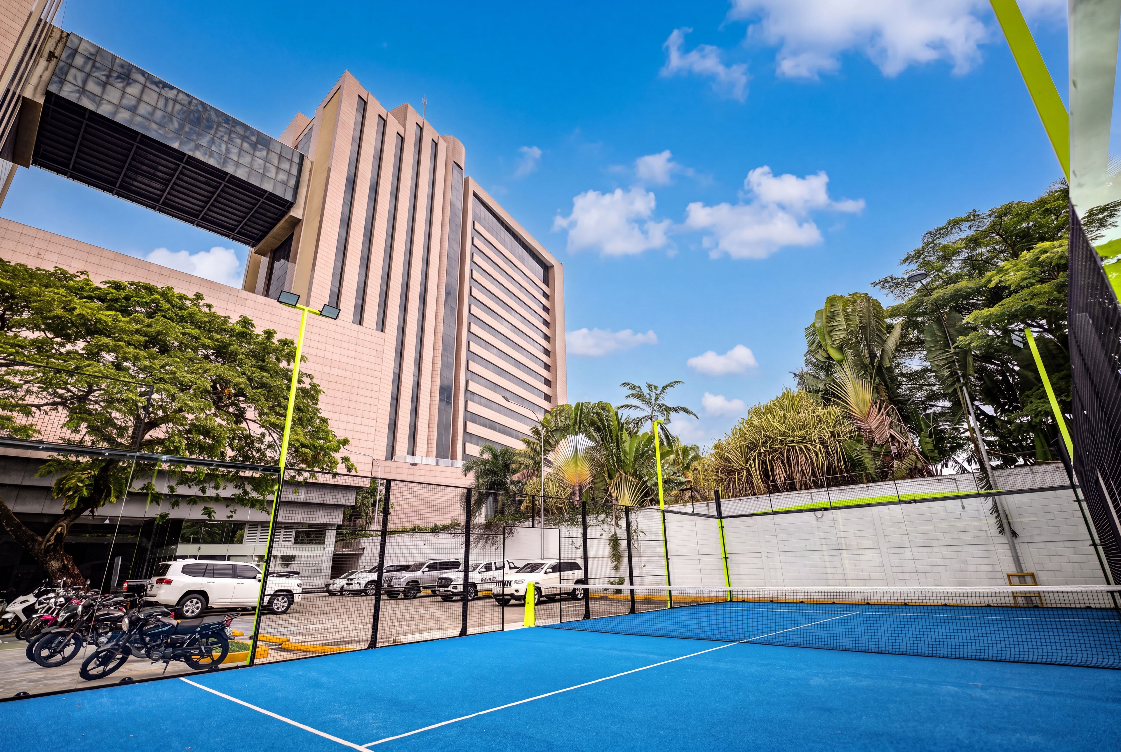 A tennis court with a net, surrounded by a fence, and a parking area with vehicles and motorcycles.