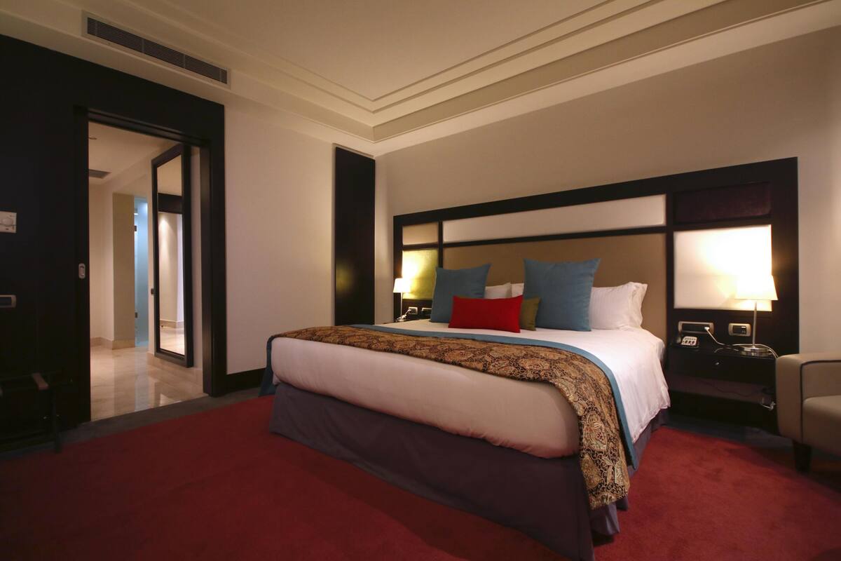 Een hotelkamer met een groot bed, een nachtkastje met een lamp, een stoel en een deur die naar een andere kamer leidt.