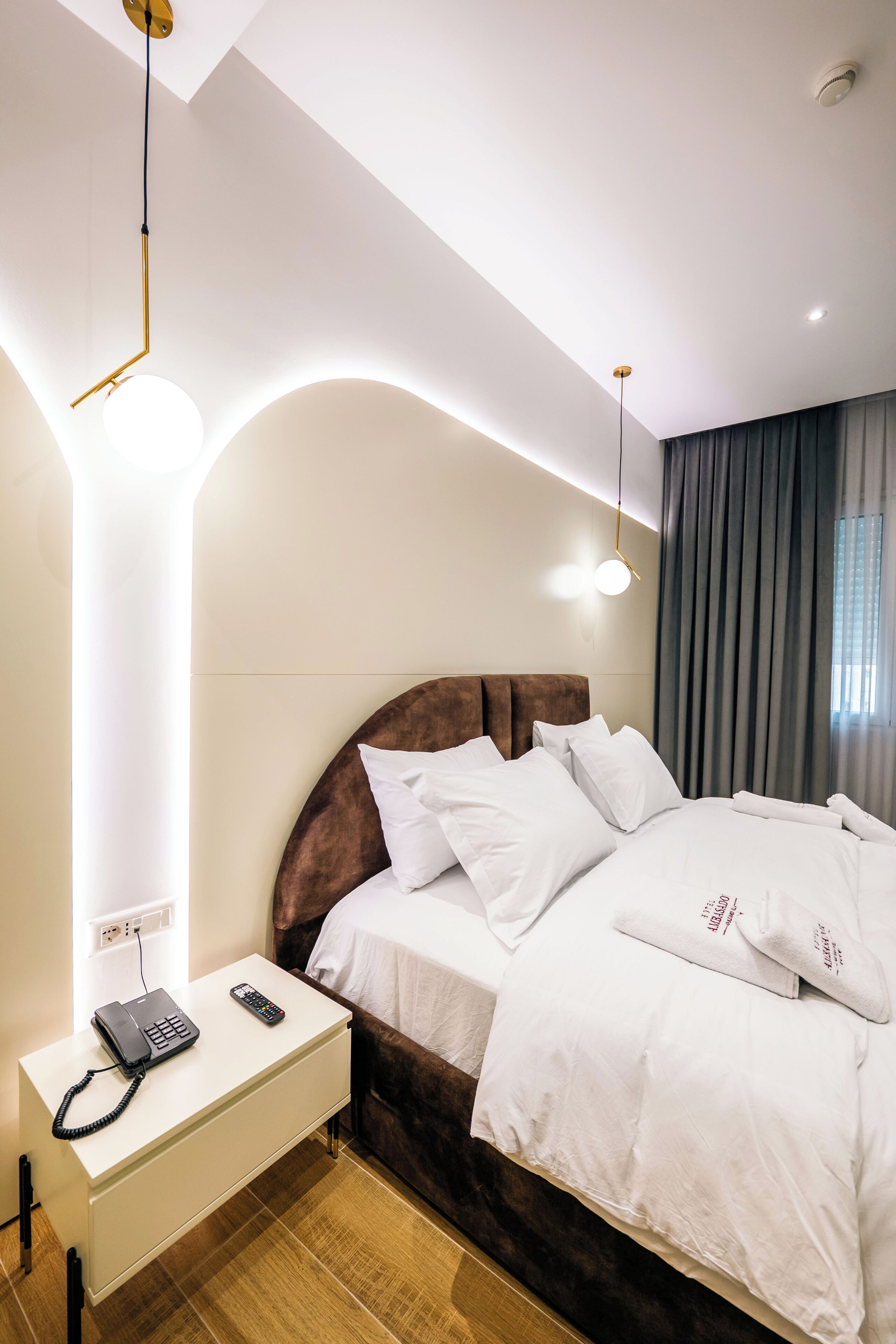 A modern hotel room with a neatly made bed, a bedside table with a phone and remote control, and a large window with curtains.
