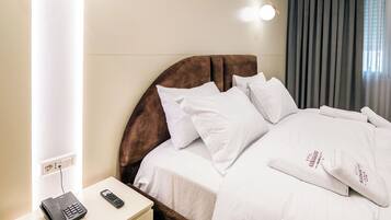 A modern hotel room with a neatly made bed, a bedside table with a phone and remote control, and a large window with curtains.