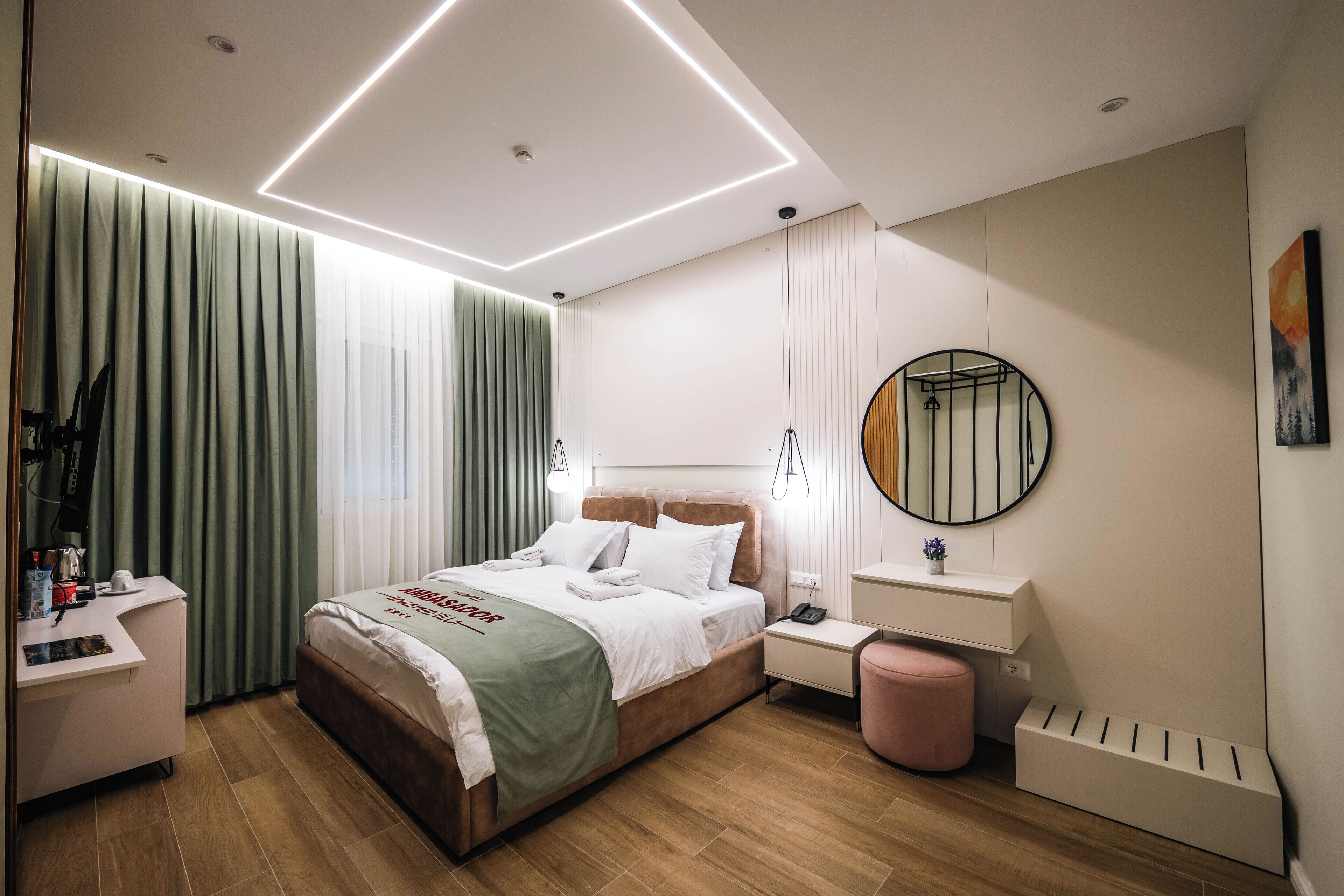 Habitació d'hotel moderna amb un llit ben fet, una taula de nit amb una làmpada, un escriptori petit amb una cadira i una finestra gran amb cortines.
