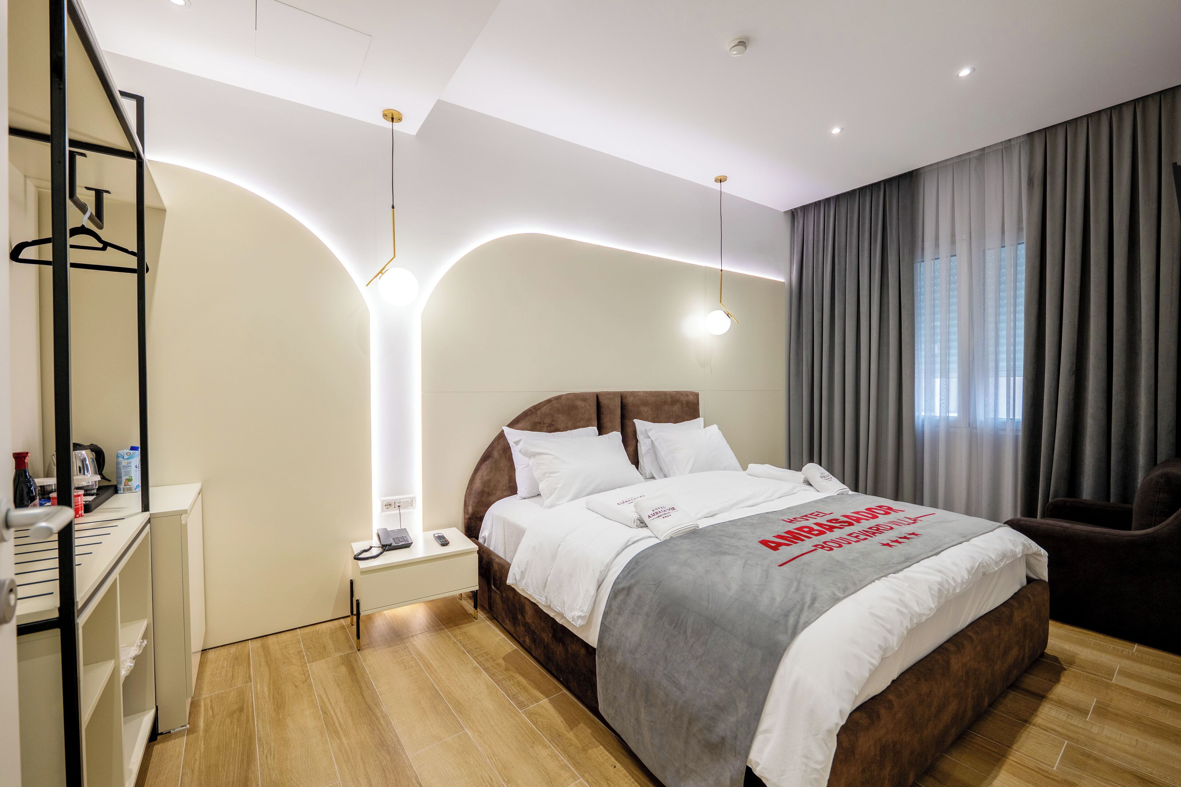 A modern hotel room with a large bed, bedside tables, a wardrobe, and a desk with a chair.