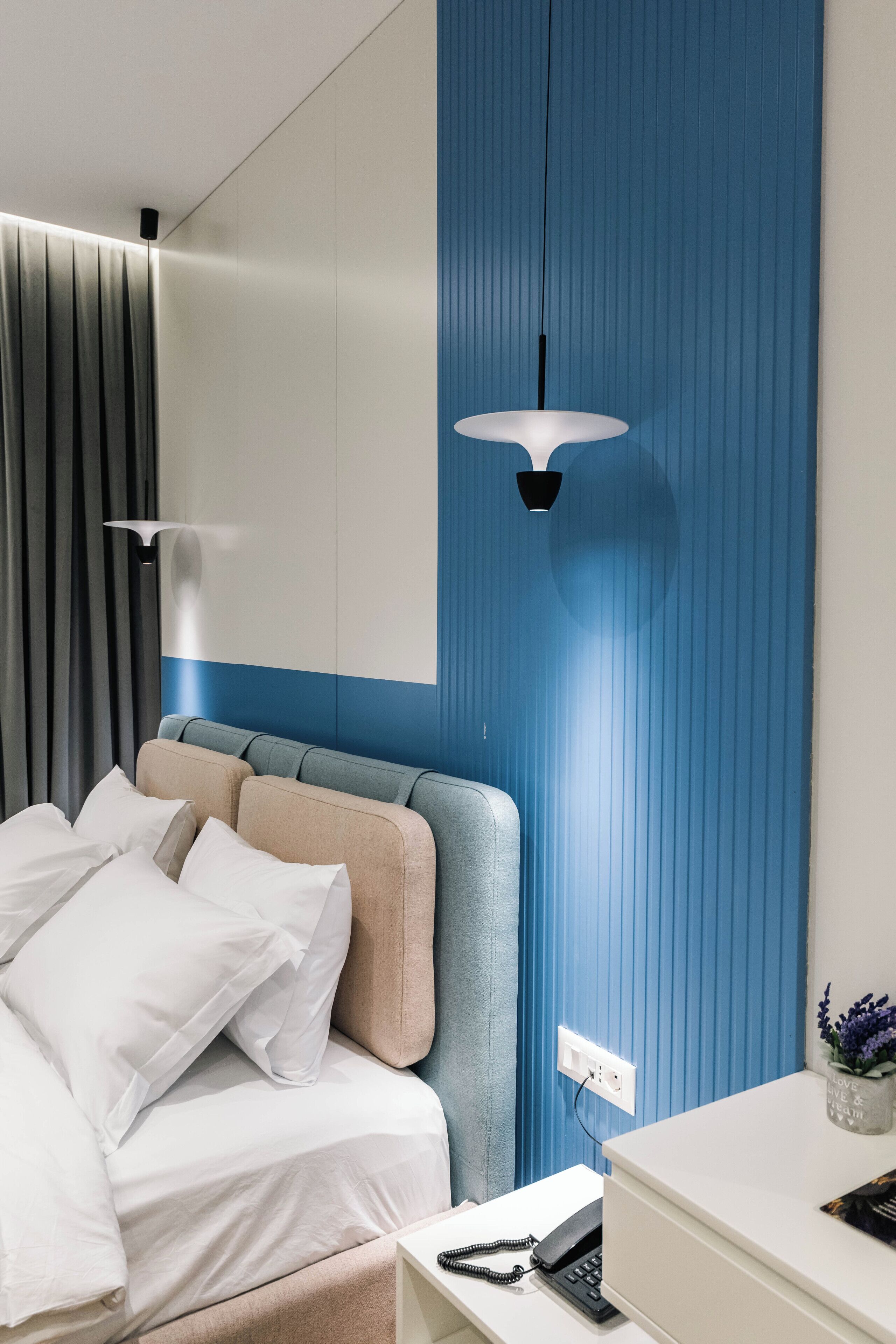 Quarto de hotel com cama, mesa de cabeceira com telefone, luminária na parede e tomada.