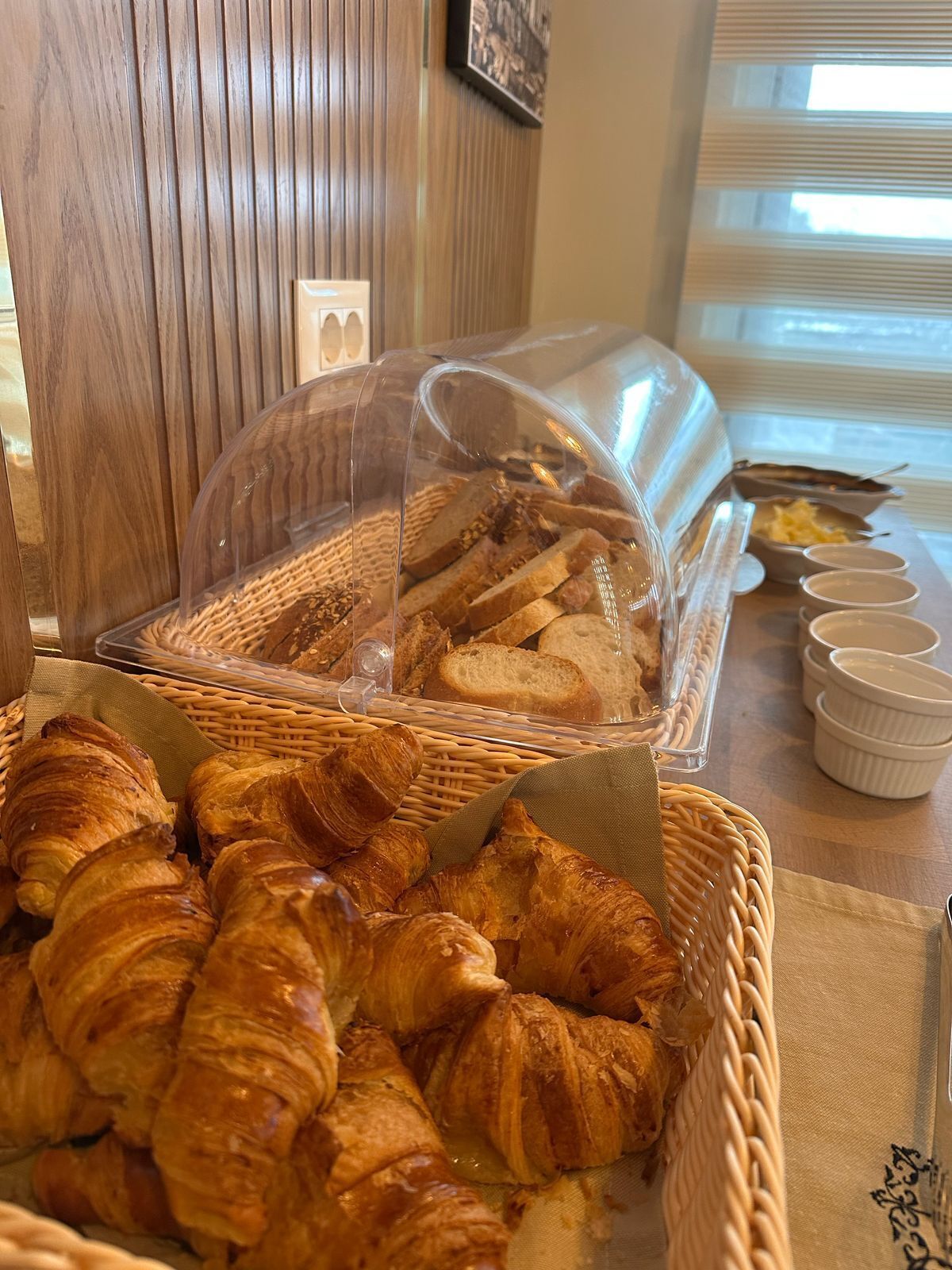 A breakfast buffet with croissants and bread, a wicker basket, and a clear plastic cover over the bread.