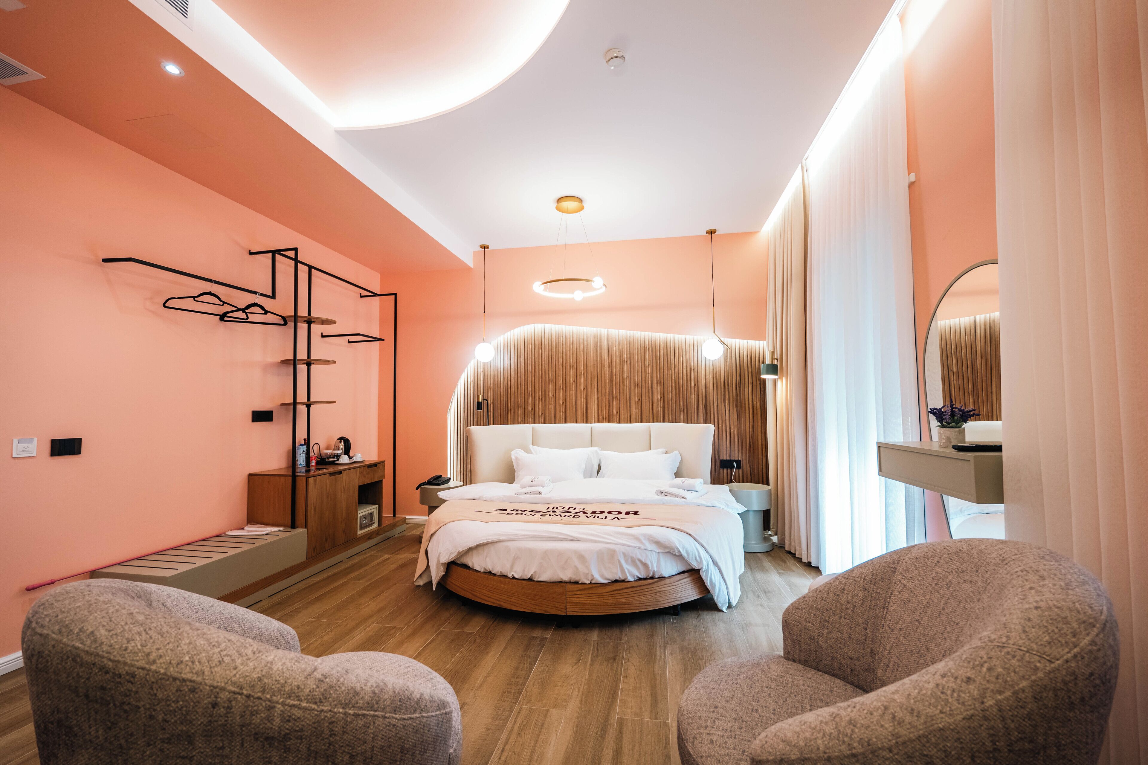 A modern hotel room with a round bed, a wooden side table, a wardrobe, and two armchairs.