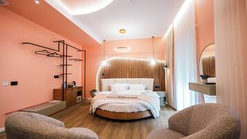 A modern hotel room with a round bed, a wooden side table, a wardrobe, and two armchairs.