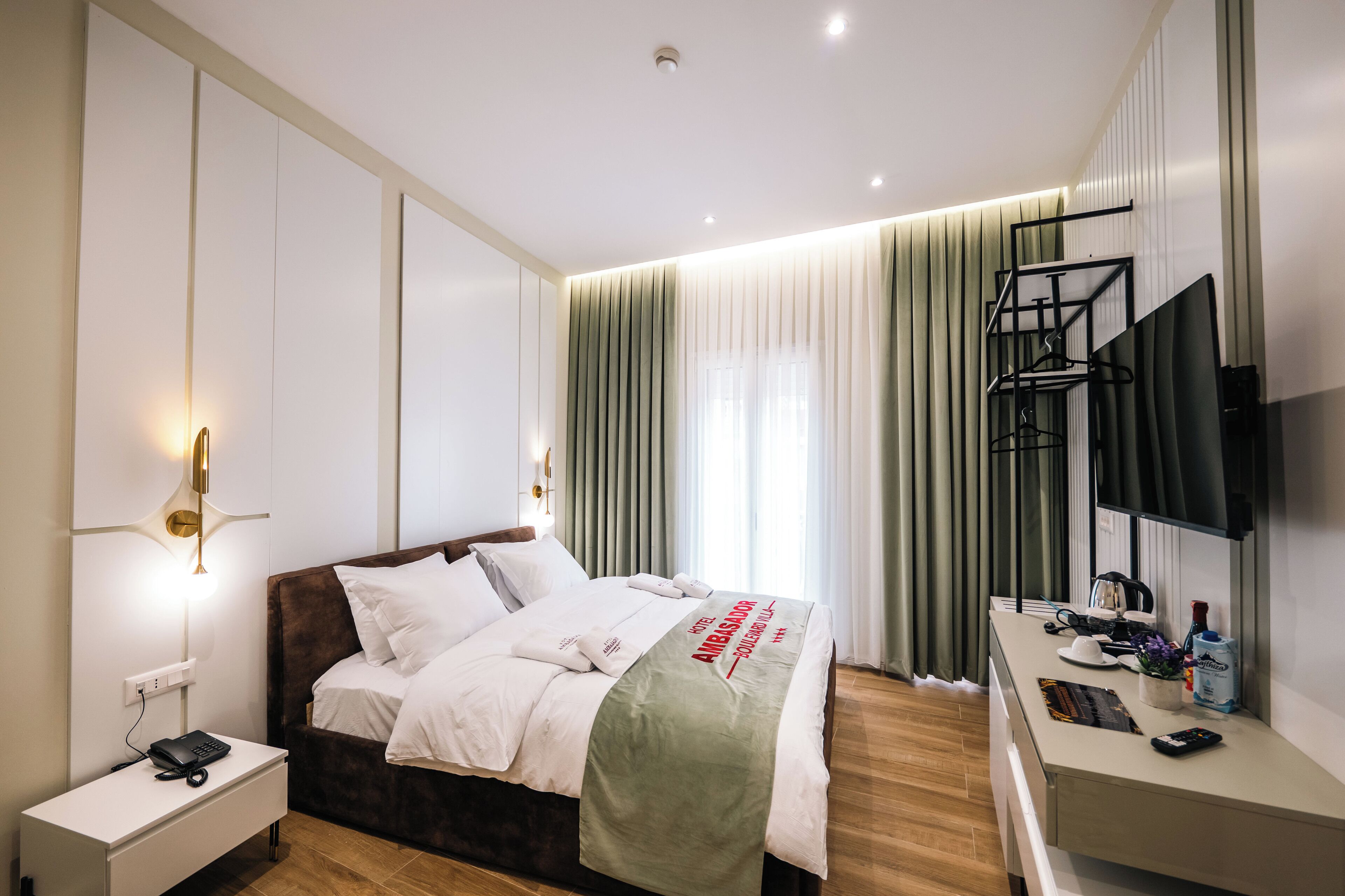 Una habitación de hotel moderna con cama, mesita de noche, lámpara, televisor y cortinas.