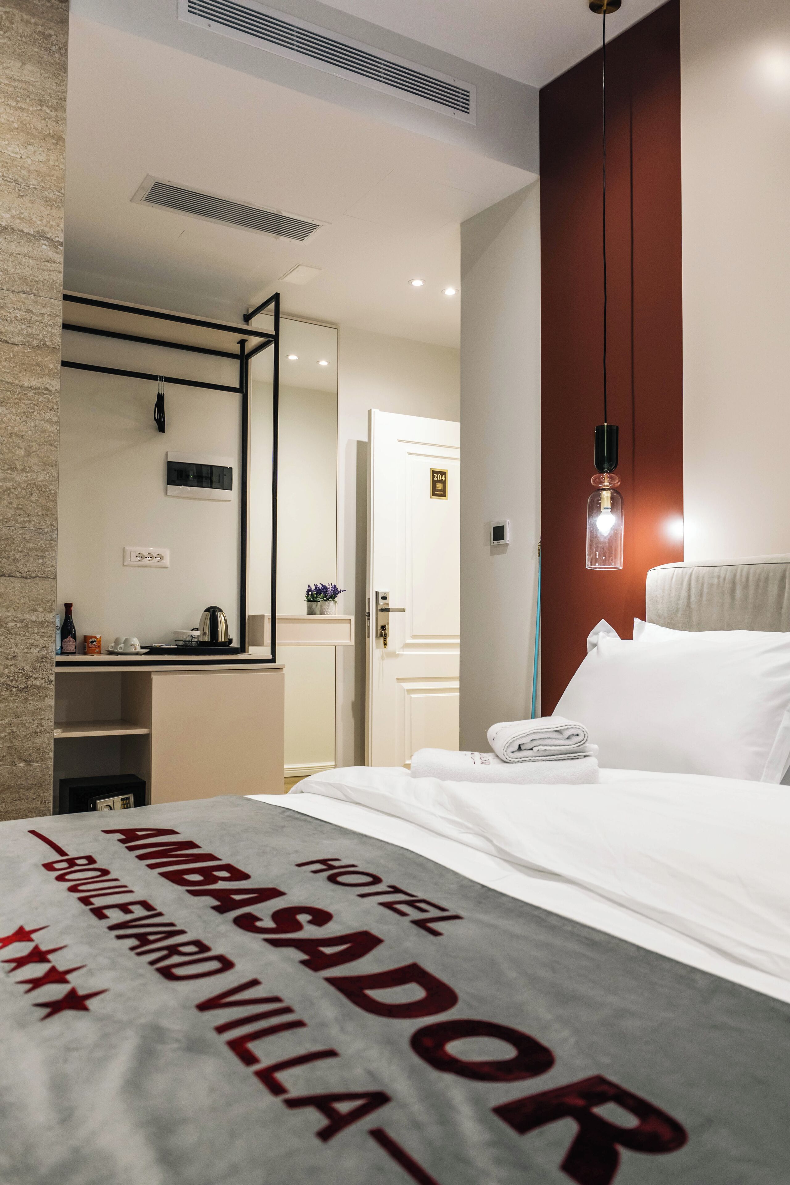 A hotel room with a bed, a desk with a kettle, a mirror, and a hanging light fixture.