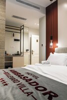 A hotel room with a bed, a desk with a kettle, a mirror, and a hanging light fixture.