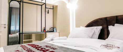 A hotel room with a bed, a desk, and a mirror. The bed has a white comforter and a pillow with the hotel name. The desk has a lamp and a bottle. The mirror has a black frame.