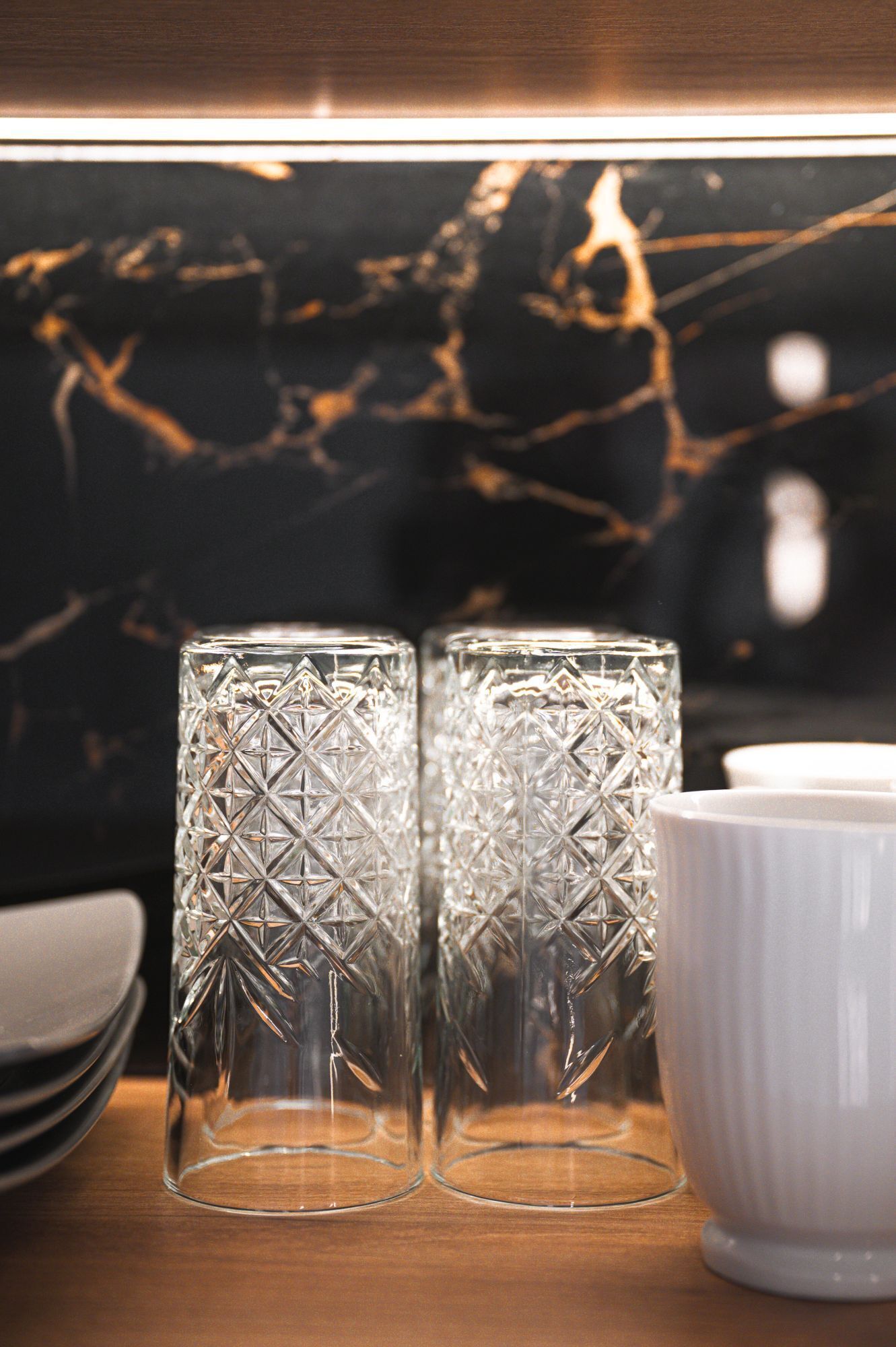 Two clear glass candle holders with geometric patterns, a white ceramic bowl, and a dark marble backsplash.