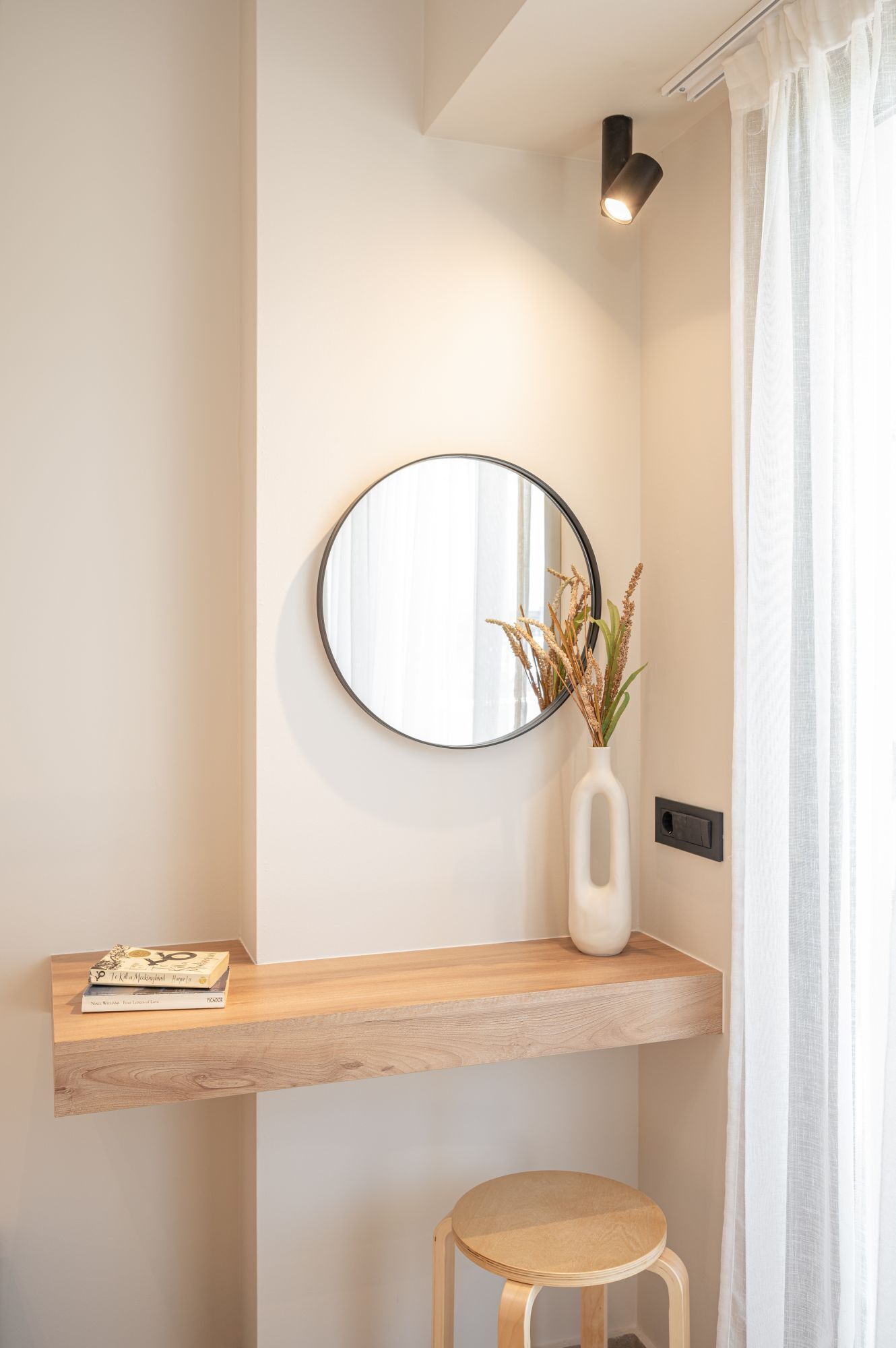 A minimalist bathroom with a wooden shelf holding books, a round mirror, and a vase with dried plants.