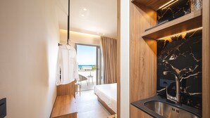 A modern hotel room with a wooden bench, a bed, a small kitchenette with a marble backsplash, and a view of the sea. - Palm Tree Beach Suites (Zakynthos)