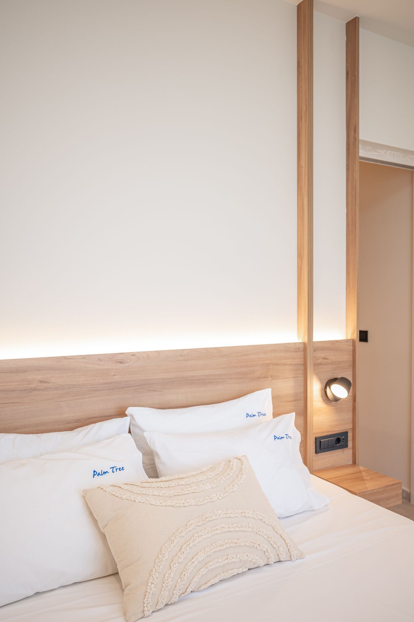 Una cama bien hecha con sábanas y almohadas blancas, con la etiqueta "Palm Tree". Se observa también un cabecero de madera y una mesita de noche con una lámpara.