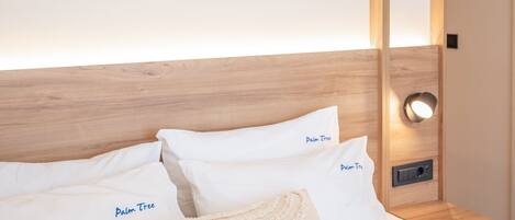 Una cama bien hecha con sábanas y almohadas blancas, con la etiqueta "Palm Tree". Se observa también un cabecero de madera y una mesita de noche con una lámpara.
