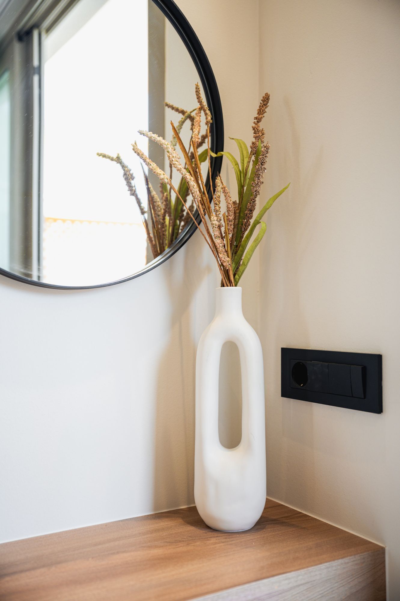 A white vase with a unique shape containing dried plants on a wooden surface next to a wall-mounted switch.