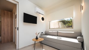 A modern living room with a grey sofa, a small wooden side table, a mounted flat-screen TV, and a window with a view of greenery. - Palm Tree Beach Suites (Zakynthos)