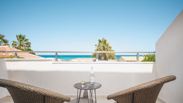 A balcony with two wicker chairs and a small table, overlooking a beach with palm trees and a clear blue sky. - Palm Tree Beach Suites (Zakynthos)