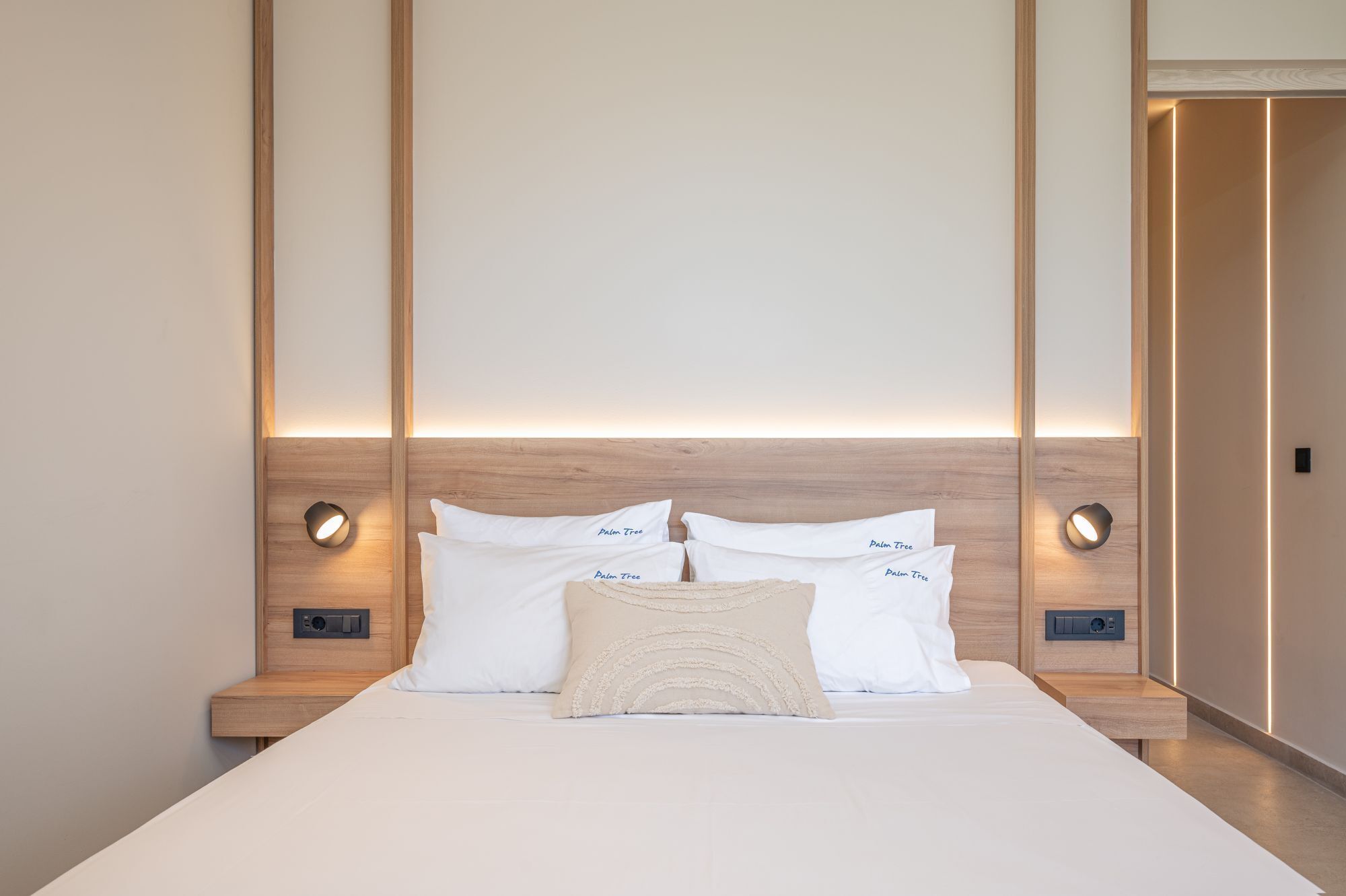 Una cama bien hecha con almohadas blancas y beige, cabecera de madera y lámparas de noche.