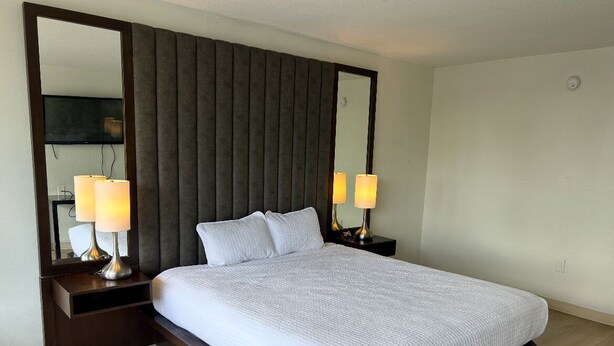 A neatly made bed with a white comforter, two bedside lamps, a large mirror, and a dark headboard. - 1st Place at Wesley (Decatur)