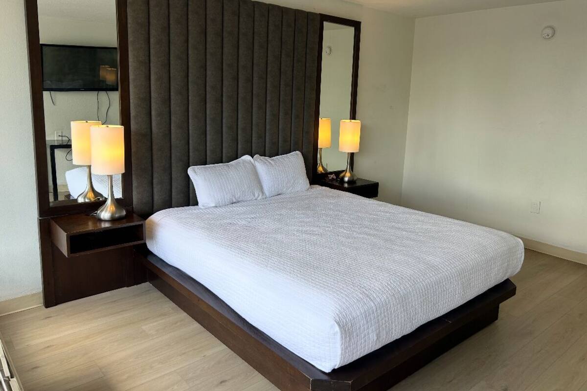 A neatly made bed with a white comforter, two bedside lamps, a large mirror, and a dark headboard.