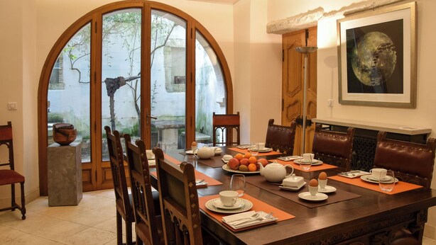 A dining area with a large wooden table set for a meal, surrounded by leather chairs, with a view through an arched window to the outside. - Chambre d'Art (Dax)