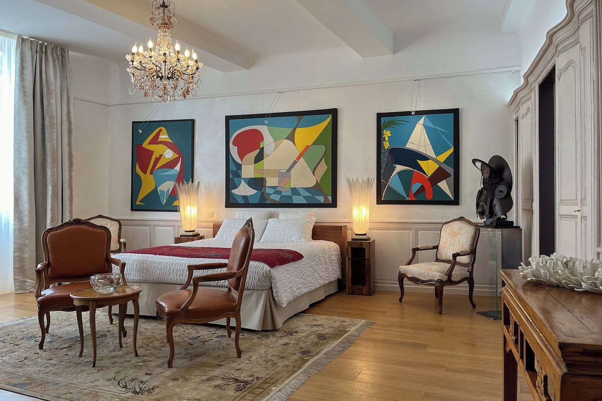 A hotel room with a bed, two chairs, a table, a chandelier, and abstract paintings on the wall.