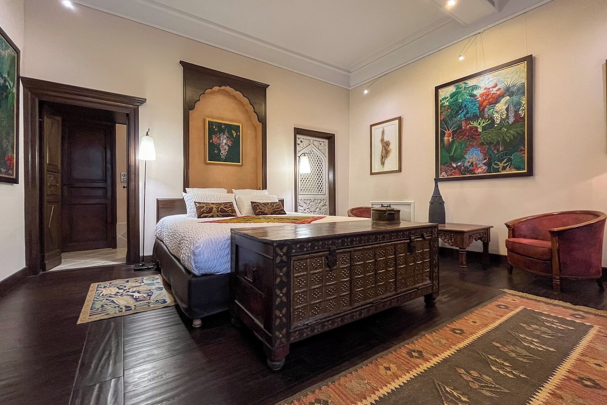 A bedroom with a large bed, a wooden chest of drawers, a red armchair, and a painting on the wall.