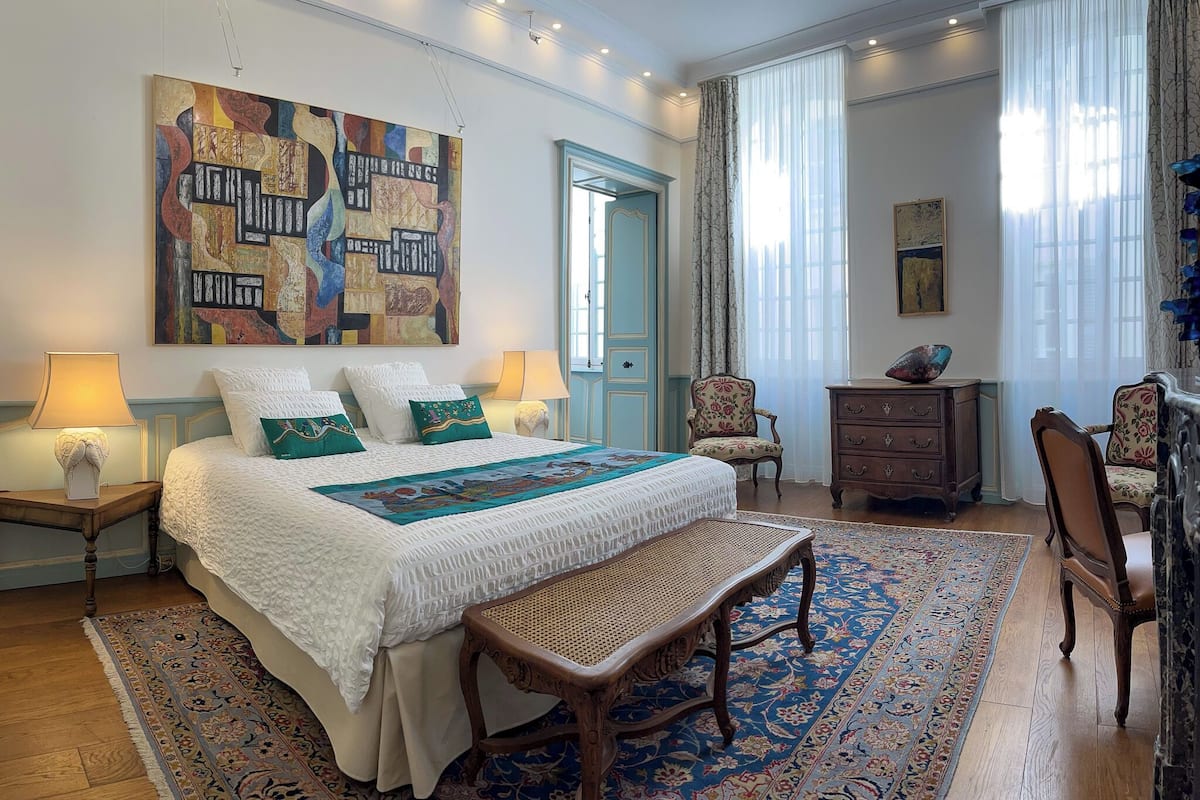 A well-lit bedroom with a large bed, a wooden bench, a dresser, and a chair.