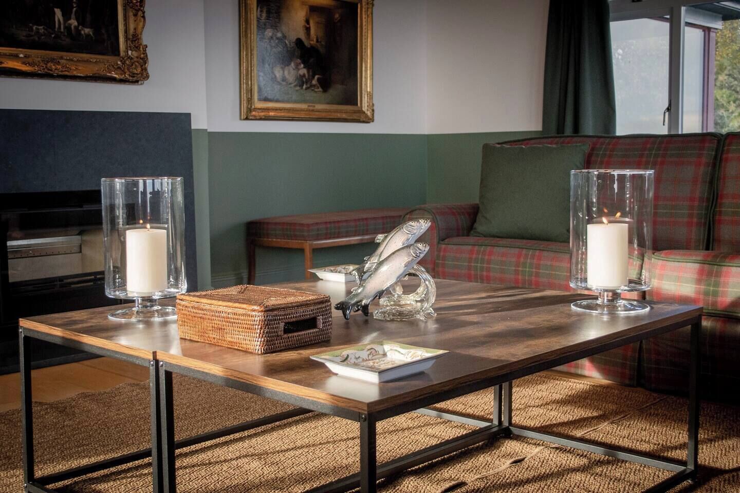 Een woonkamer met een houten salontafel, twee kandelaars van glas met brandende kaarsen, een geweven mand en een decoratief beeld van een vis.