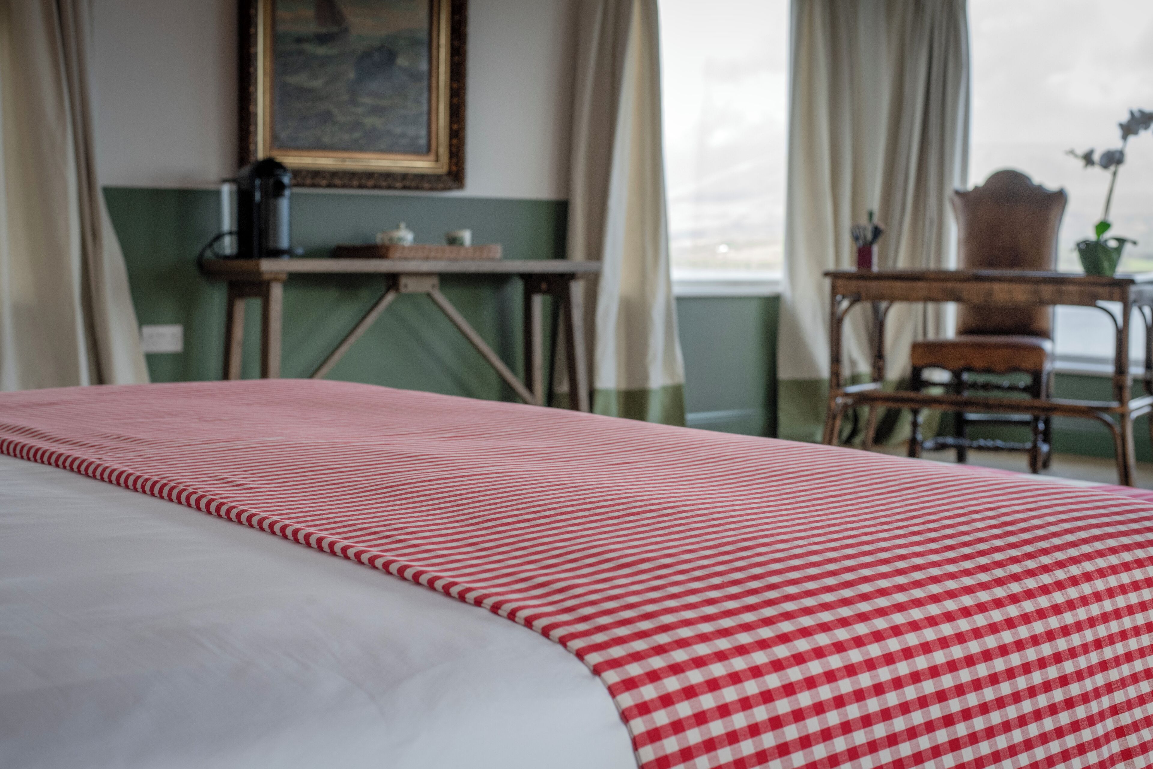Una habitación con una cama cubierta por un edredón rojo y blanco a cuadros, una silla de madera, una mesita con un jarrón de flores y un cuadro en la pared.