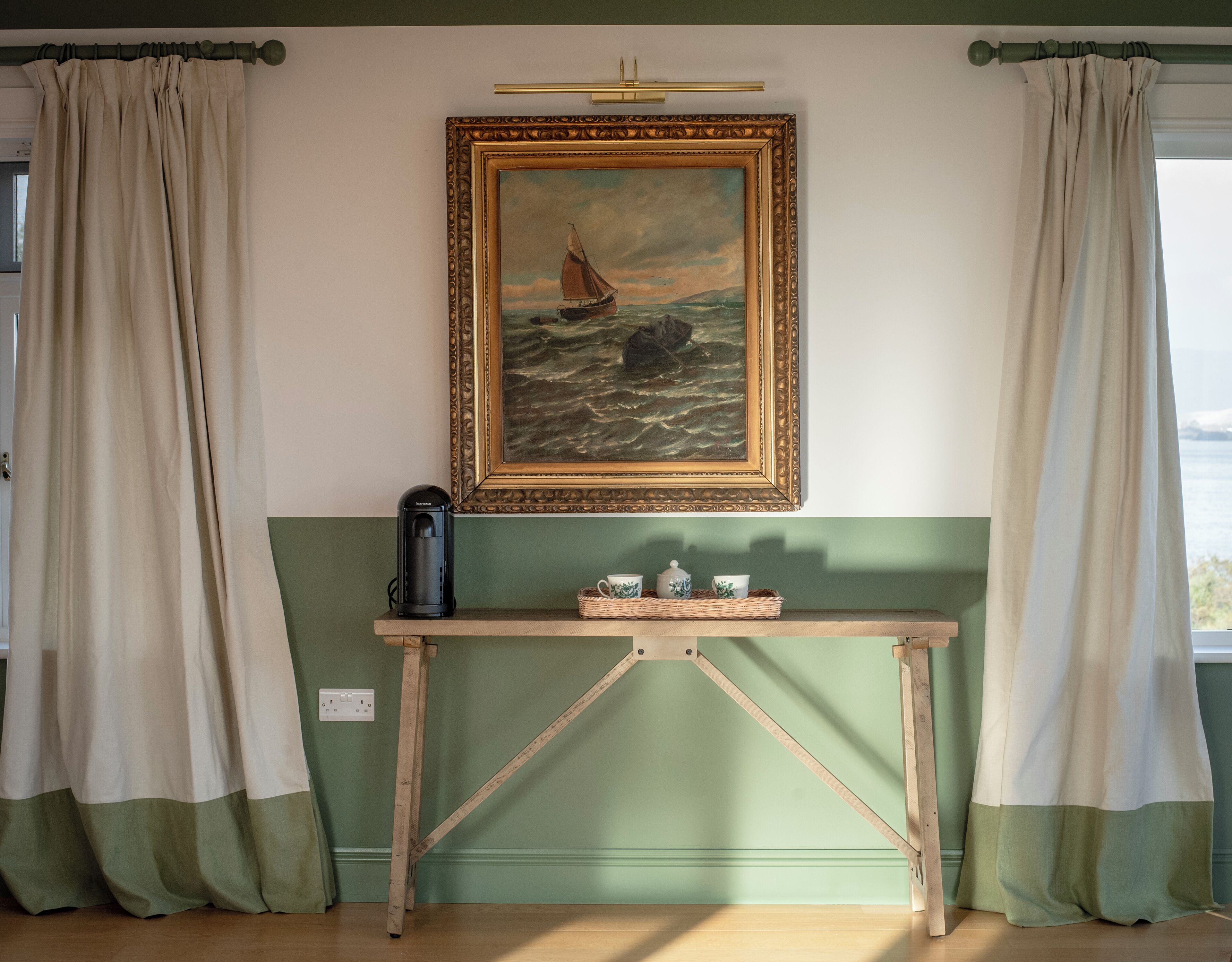 Una stanza con un quadro raffigurante una barca a vela su un mare calmo, una macchina per il caffè e un vassoio con tazze e piattini su un tavolo in legno.