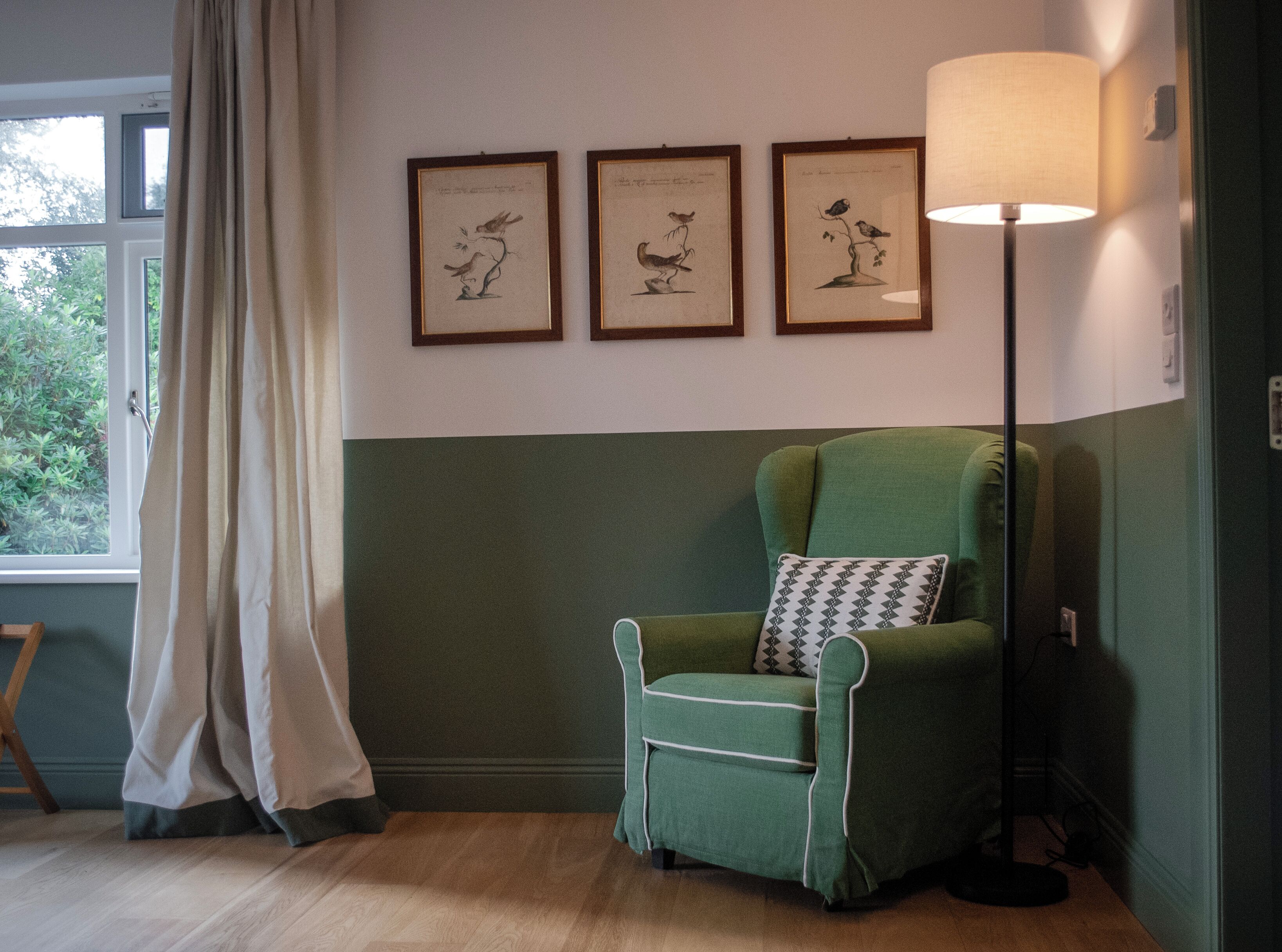 Ein Zimmer mit einem grünen Sessel, einer Stehlampe und drei gerahmten Pflanzenbildern an der Wand.